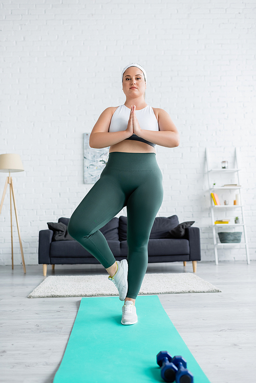 Plus size woman standing with praying hands on fitness mat near dumbbells at home