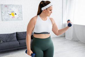 Side view of plus size woman training with dumbbells in living room
