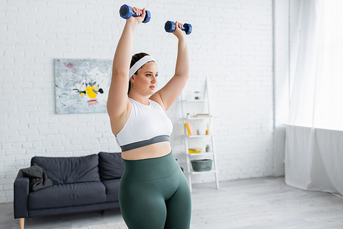 Plus size woman in sportswear training with dumbbells in living room