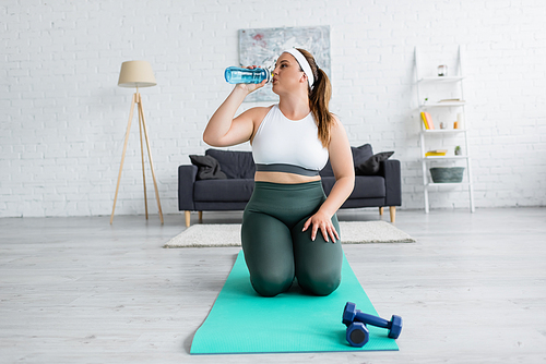 Plus size woman in sportswear drinking water near fitness mat and dumbbells at home