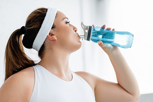 Side view of plus size woman in sportswear drinking water at home