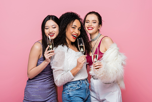 Positive multicultural women in festive clothes holding glasses of champagne isolated on pink
