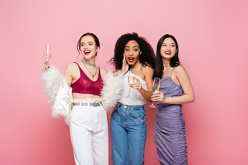 Excited african american woman holding champagne near cheerful interracial friends isolated on pink
