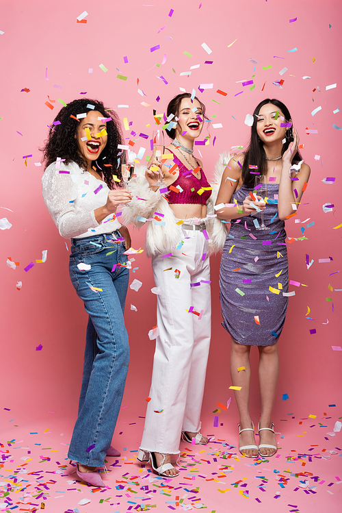 Pretty multicultural women with champagne  under confetti on pink background