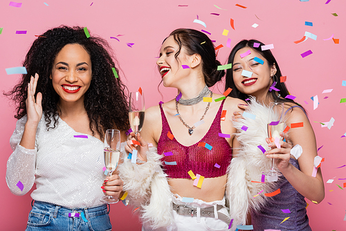 Trendy multiethnic women with champagne standing under confetti on pink background