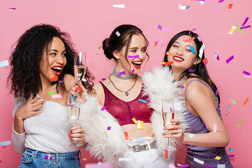 Stylish multiethnic women holding glasses of champagne under confetti on pink background