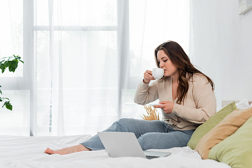 Young body positive woman drinking coffee near laptop on bed at home