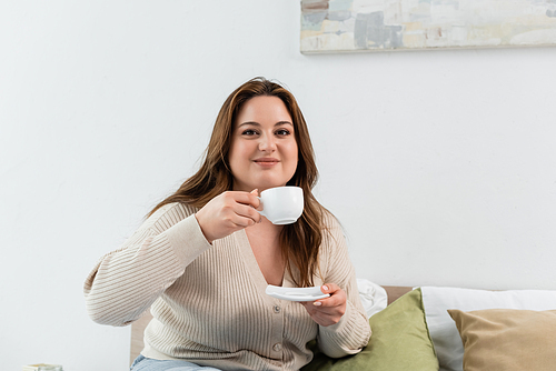 Pleased body positive woman holding cup and saucer on bed at home