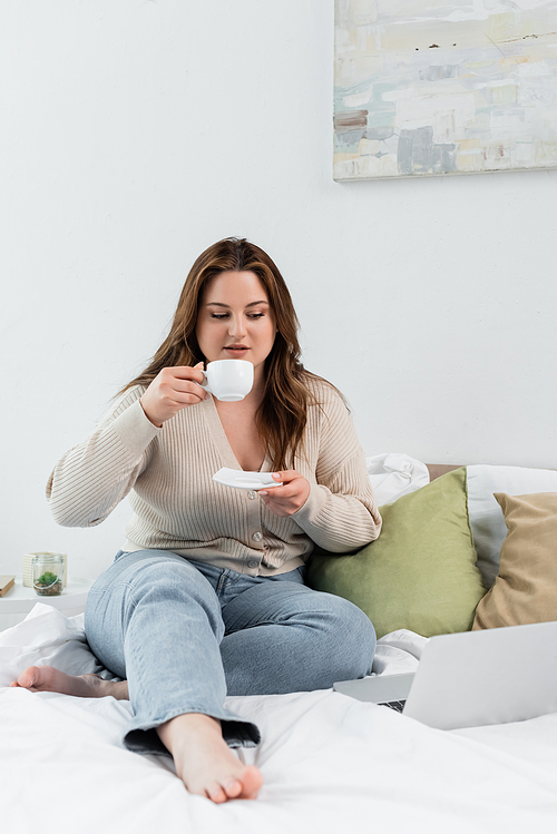 Young woman with overweight holding cup of coffee near laptop on bed