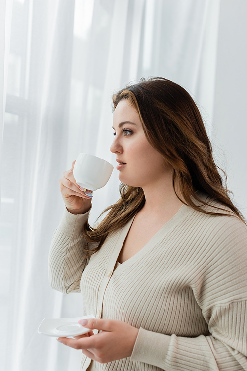 Young plus size woman holding cup of coffee near curtains at home