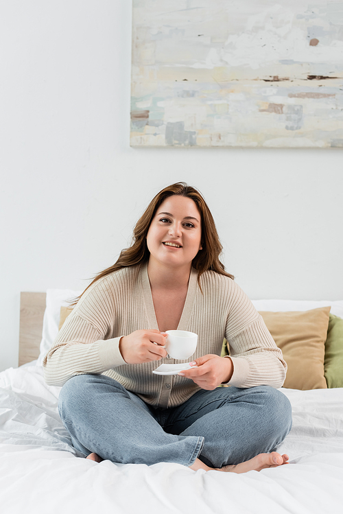 Happy plus size woman holding cup of coffee on bed