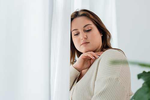 Portrait of pretty body positive woman standing near blurred curtains at home