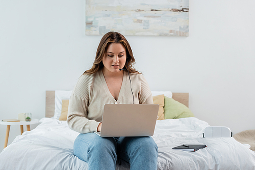 Freelancer with overweight using laptop and headset near notebook on bed