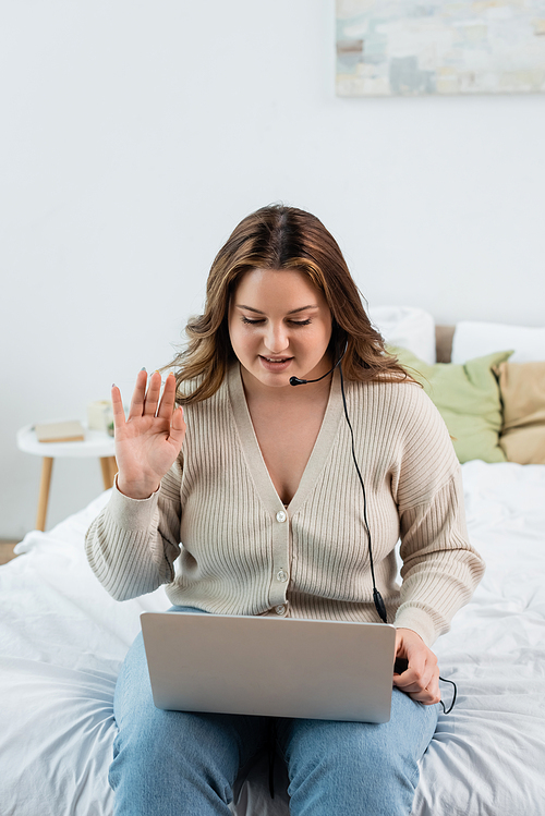 Plus size freelancer in headset having video call on laptop in bedroom