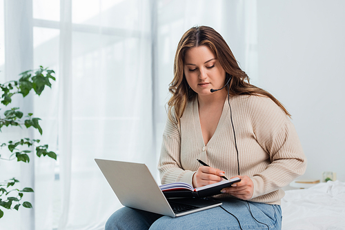 Young freelancer with overweight writing on notebook while using laptop and headset in bedroom