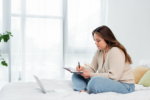 Young body positive freelancer in headset writing on notebook near laptop in bedroom