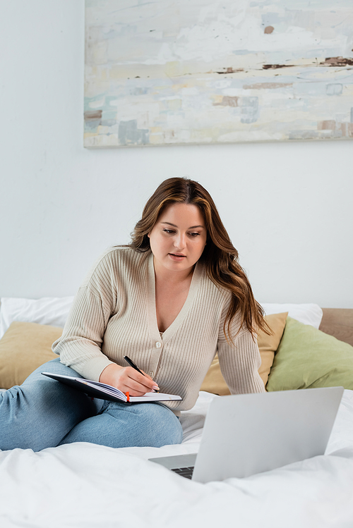 Brunette woman with overweight writing on notebook near blurred laptop on bed