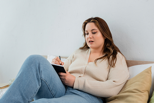 Young plus size woman writing on notebook on bed