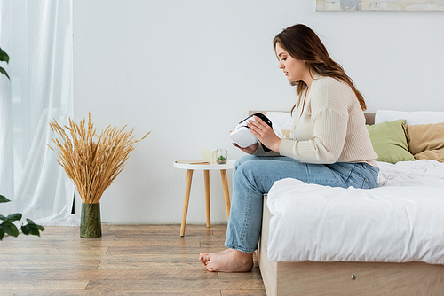 Side view of young body positive woman holding vr headset on bed at home