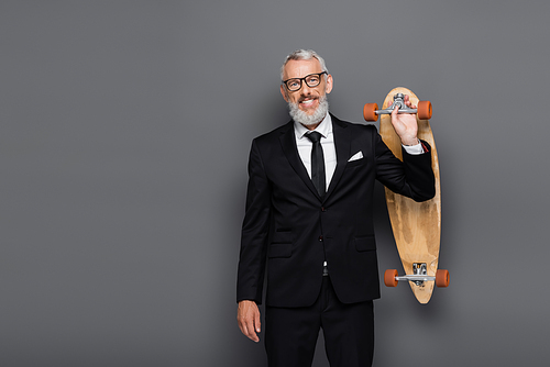 joyful middle aged businessman in suit and glasses holding longboard on grey