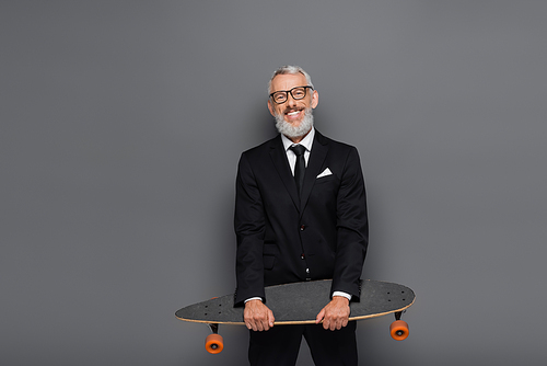 happy middle aged businessman in suit and glasses holding longboard on grey