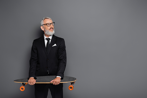 bearded middle aged businessman in suit and glasses holding longboard on grey