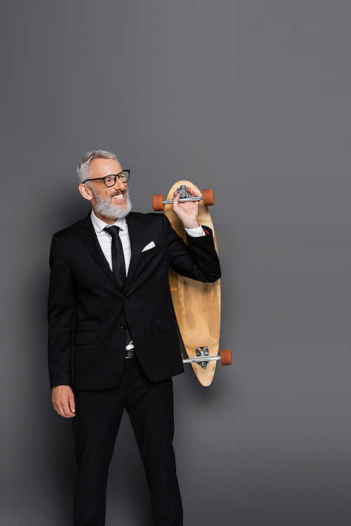 cheerful middle aged businessman in suit and glasses holding longboard on grey