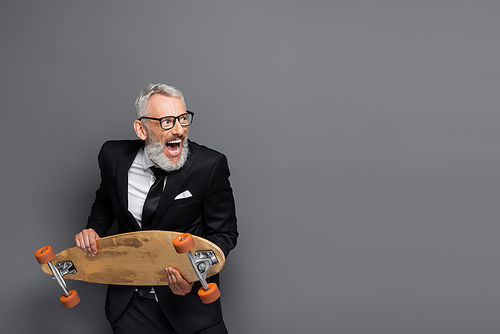 middle aged businessman in suit and glasses holding longboard and screaming on grey