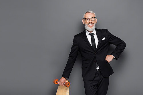 happy middle aged businessman in suit and glasses holding longboard while posing with hand on hip on grey