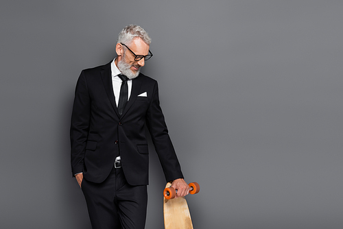 smiling middle aged businessman in suit and glasses holding longboard on grey