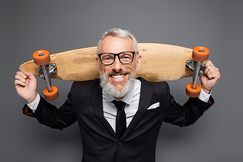 positive middle aged businessman in suit and glasses holding longboard on grey