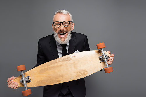 joyful and mature businessman in suit and glasses holding longboard isolated on grey