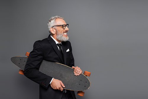 joyful middle aged businessman in suit and glasses holding longboard on grey