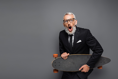 shocked middle aged businessman in suit and glasses holding longboard isolated on grey