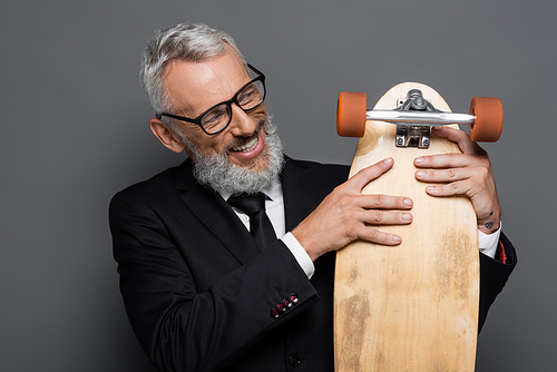 happy and mature businessman in glasses holding longboard on grey