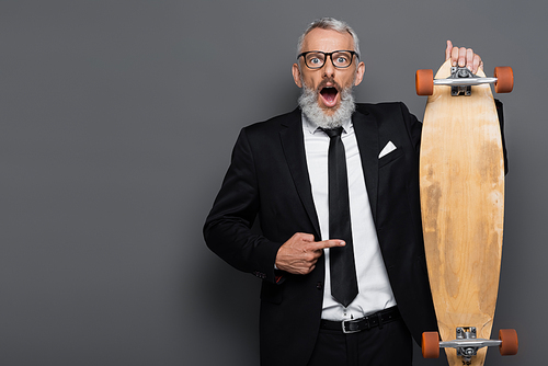 shocked middle aged businessman in suit and glasses pointing at longboard on grey
