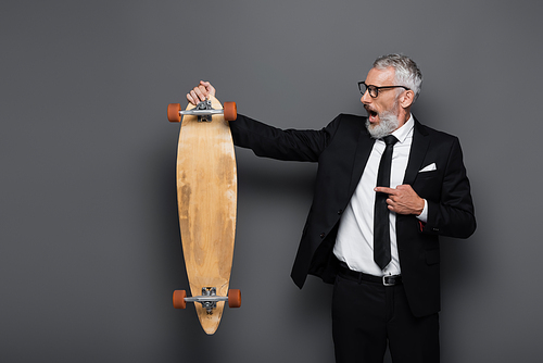 shocked and mature businessman in suit and glasses pointing at longboard on grey
