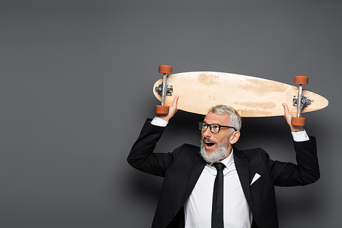 amazed and mature businessman in suit and glasses holding longboard above head on grey