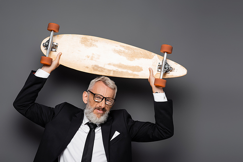 joyful and mature businessman in suit and glasses holding longboard above head on grey