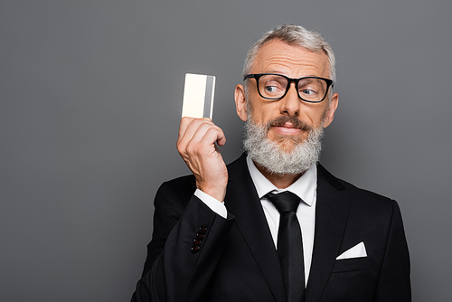 joyful and mature businessman in suit and glasses holding credit card isolated on grey