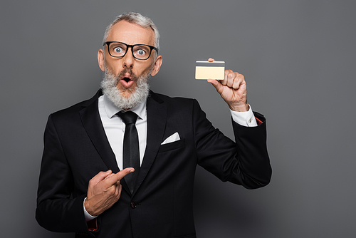 shocked and mature businessman in suit and glasses pointing at credit card isolated on grey