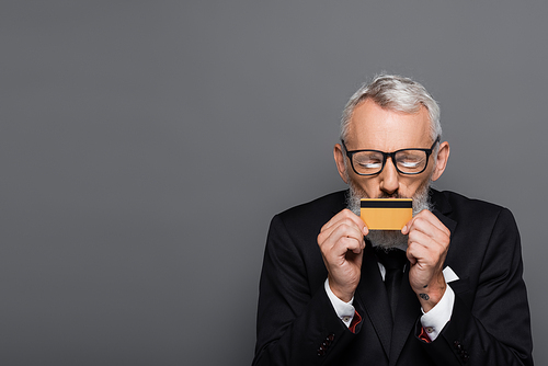 mature businessman in glasses kissing credit card isolated on grey