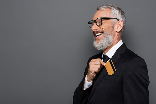 happy middle aged businessman in suit and glasses putting credit card in pocket isolated on grey