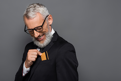 cheerful middle aged businessman in suit and glasses putting credit card in pocket isolated on grey