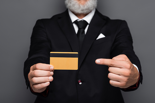 cropped view of bearded middle aged businessman in suit pointing at credit card isolated on grey