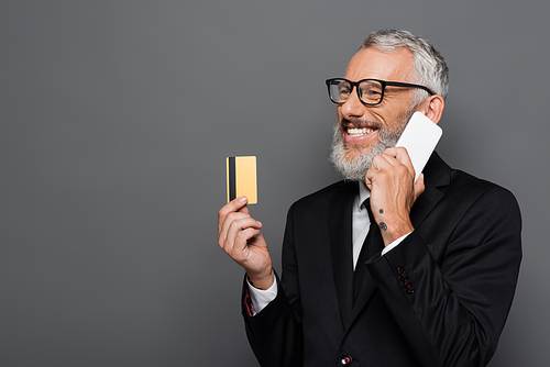 happy middle aged businessman holding credit card while talking on smartphone isolated on grey