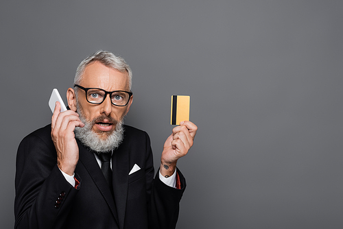surprised middle aged businessman holding credit card while talking on smartphone isolated on grey
