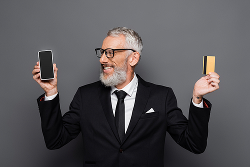 happy middle aged businessman holding credit card and smartphone with blank screen on grey