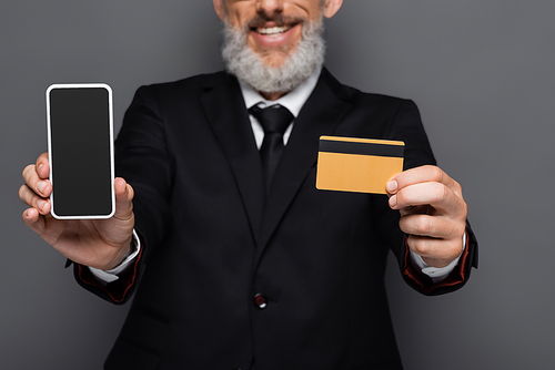cropped view of happy middle aged businessman holding credit card and smartphone with blank screen isolated on grey