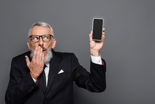 middle aged businessman holding cellphone with blank screen and covering mouth on grey